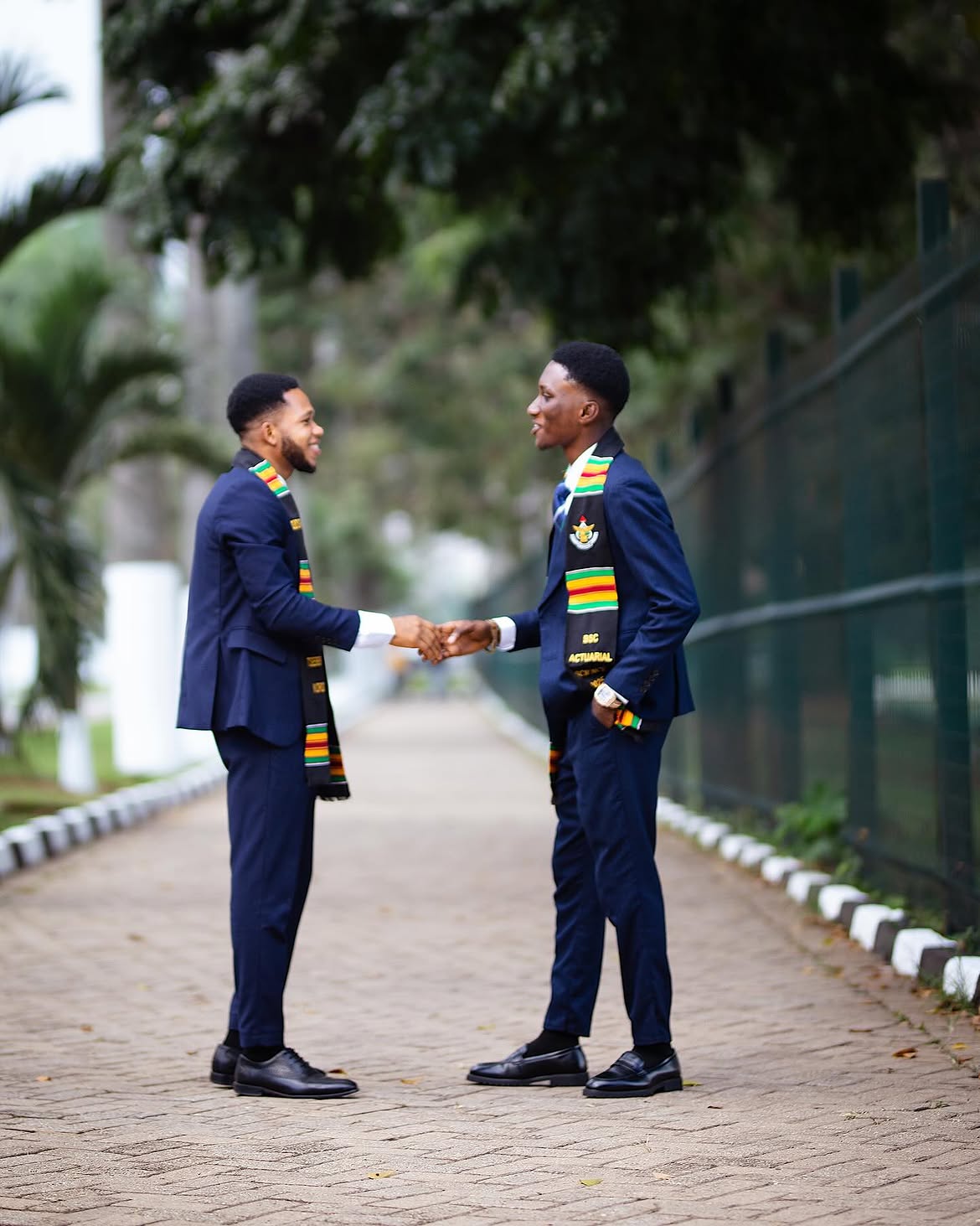 Two male graduates in navy suits and kente stoles shaking hands on a brick campus path