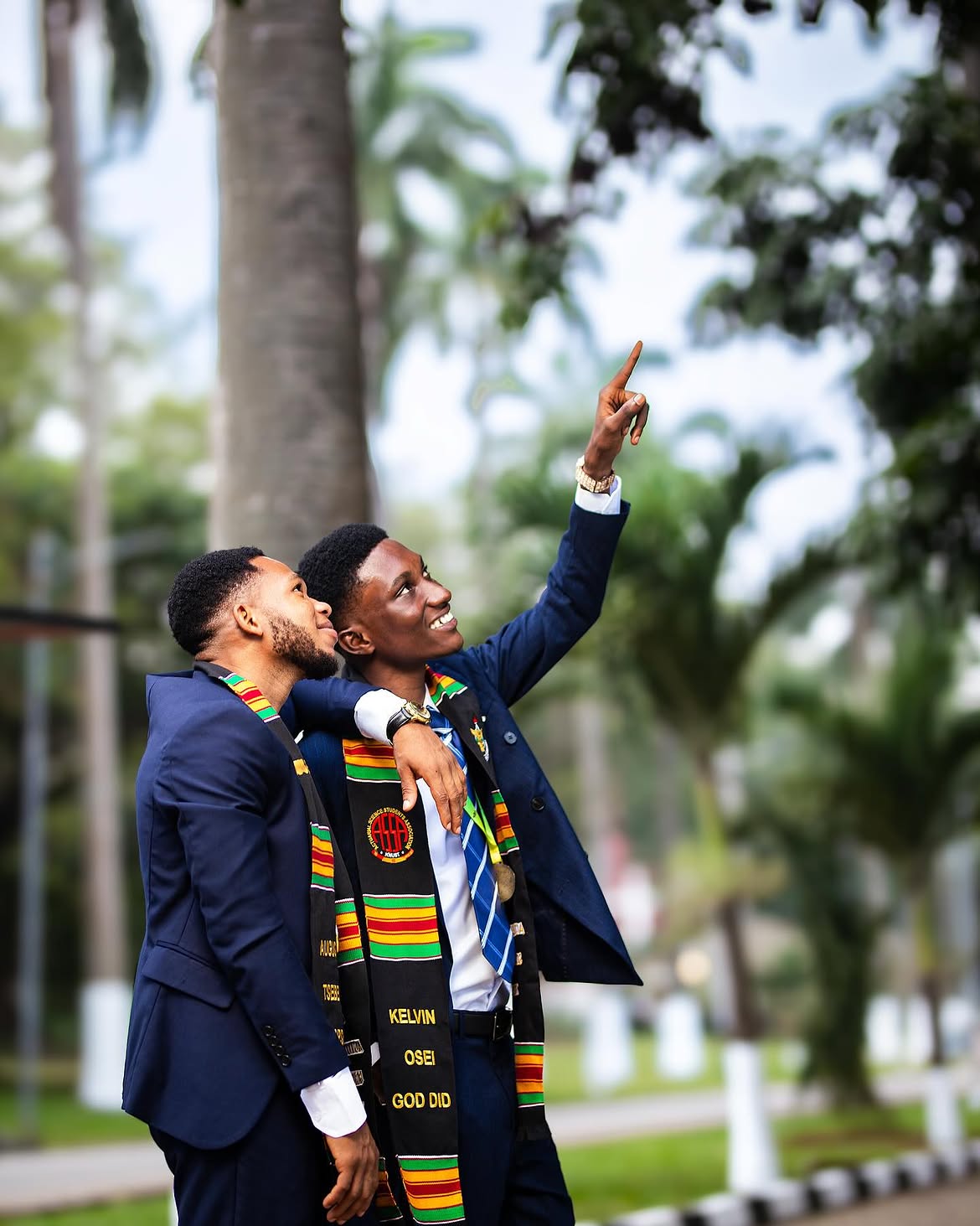 Two Ghanaian graduates in navy suits, arm around each other, one pointing skyward — brothers celebrating