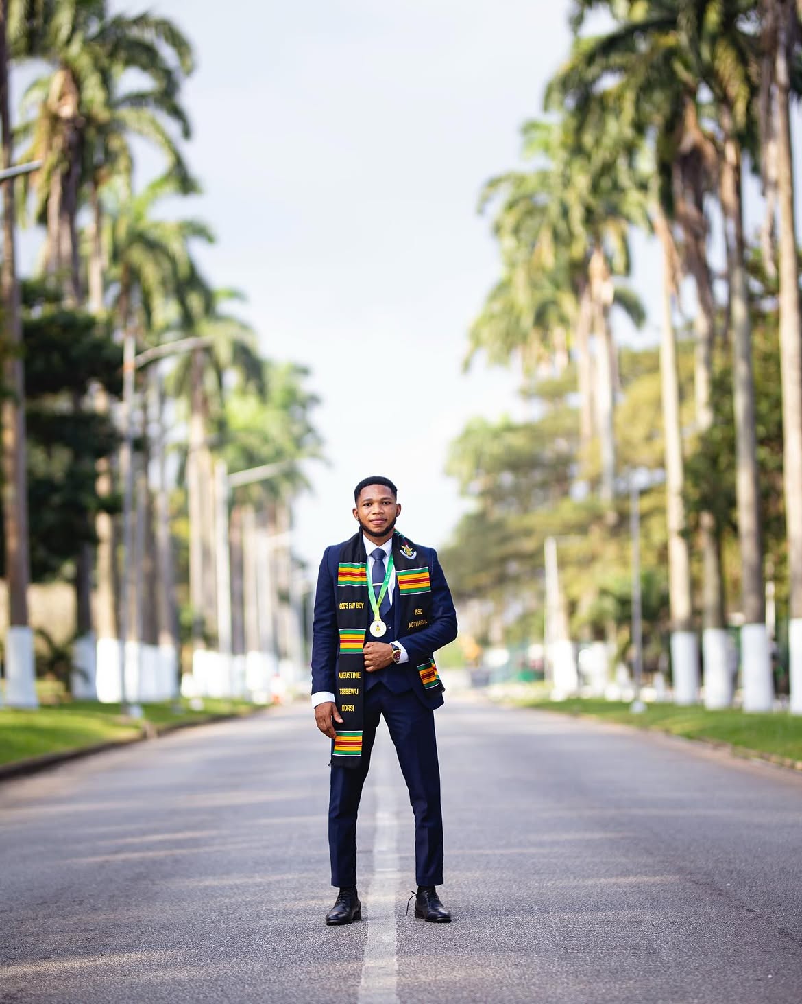 Male graduate standing composed on a long palm-lined campus road — styled portrait, editorial direction