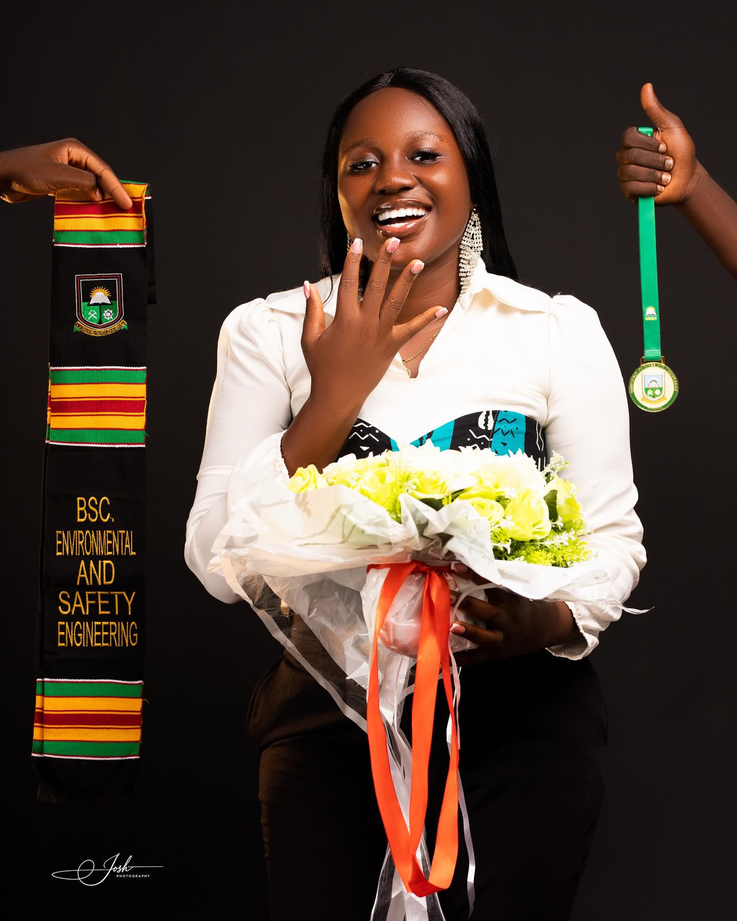 Graduate smiling joyfully in studio holding a bouquet of flowers, kente stole and medal displayed
