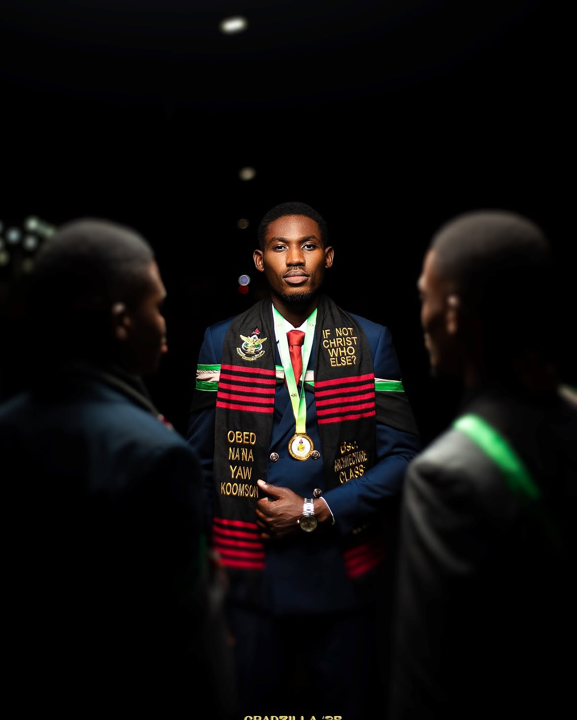Graduate standing in dramatic low-key studio lighting, flanked by silhouettes of classmates