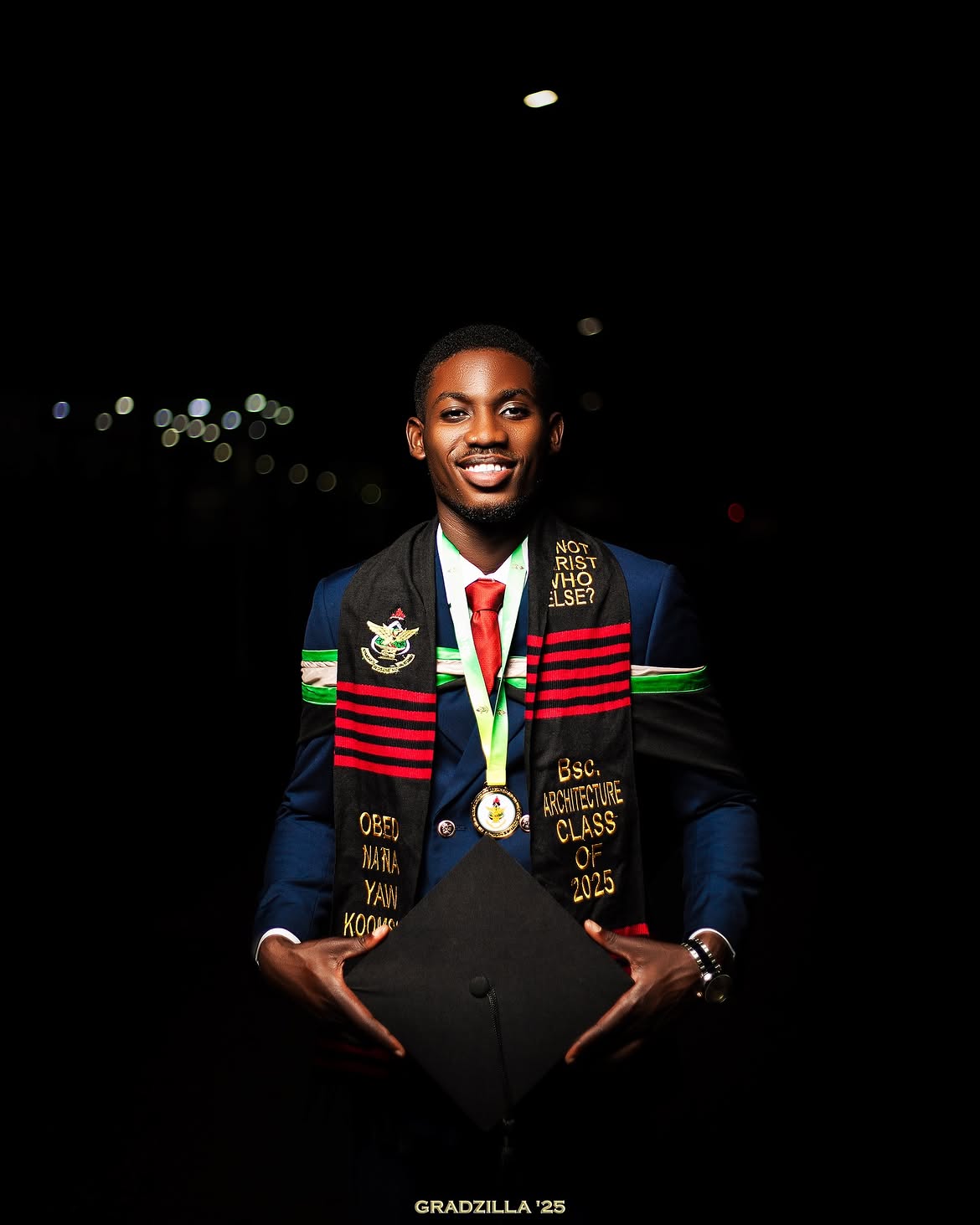 Graduate Obed Koomson smiling, holding his mortarboard against a dark night background