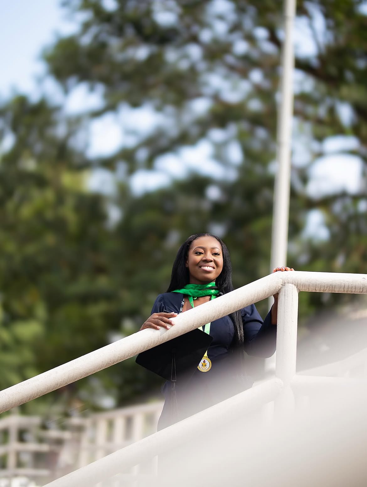 Female graduate leaning on white campus staircase railings, looking upward — the anticipation before it all begins